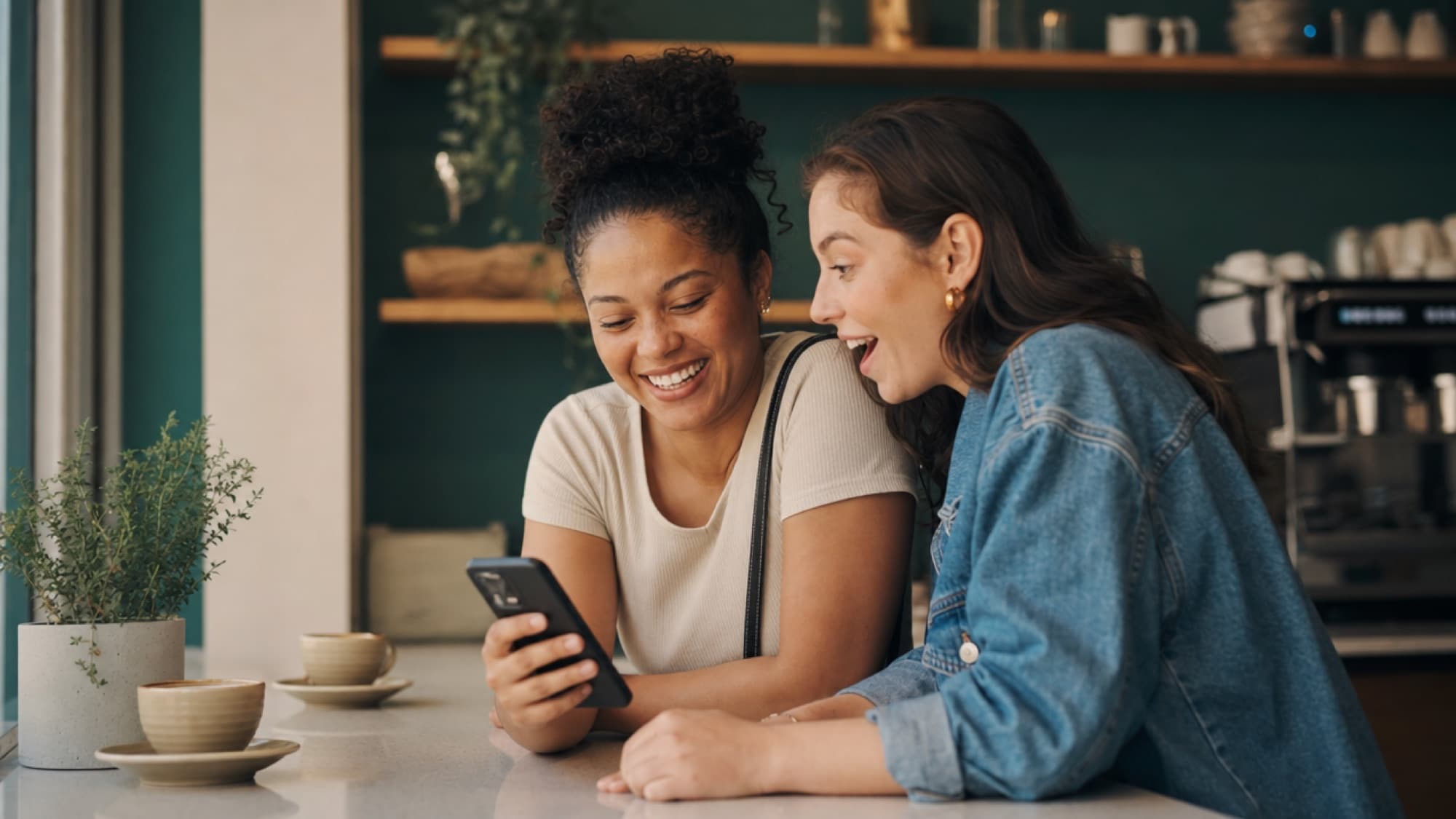Two friends at a cafe counter sharing something on a phone, both lighting up